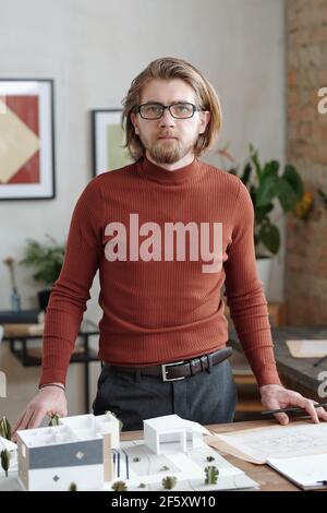 Young elegant male architect in smart casualwear standing by workplace with model of new house and yard while looking at you in office Stock Photo