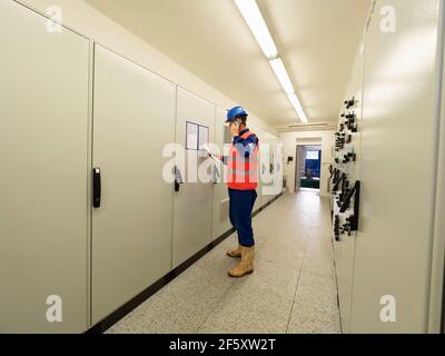 Expert in reflective vest stay in front of switch board, reading data from document and call colleague. Employee in technical corridor with control pa Stock Photo