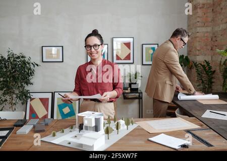 Young smiling female architect in casualwear and eyeglasses standing by table with model of new house and yard against mature male colleague Stock Photo