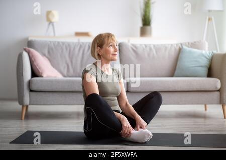 Active mature lady doing yoga, sitting in butterfly asana on sports mat at home, empty space Stock Photo
