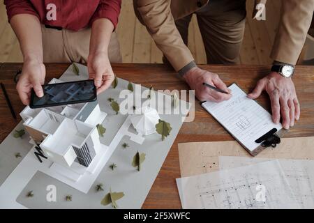 Hands of young female architect taking photo of model of new house and yard while her mature male colleague making notes below sketch on paper Stock Photo
