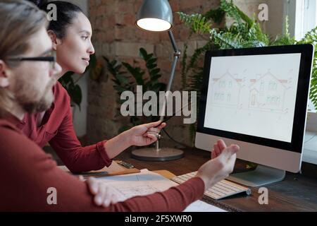 Young female architect and her male colleague discussing new project in front of computer while woman pointing at sketch of house on screen Stock Photo