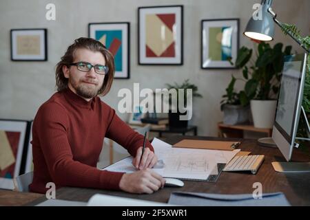 Happy young bearded male architect in eyeglasses and casualwear looking at you while sitting by table in front of camera and working over sketch Stock Photo