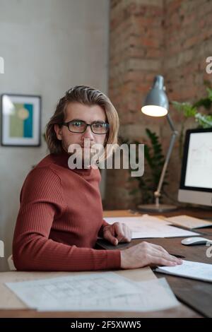 Young bearded male architect in eyeglasses and casualwear looking at you while sitting by table in front of camera and working over new project Stock Photo