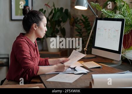 Young serious female architect in casualwear looking at sketch on paper while sitting by table in front of computer screen with picture of house Stock Photo