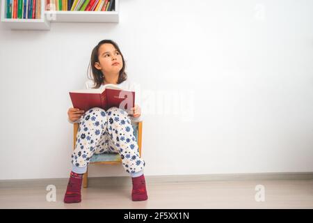 Happy smart schoolgirl reading Bible in next to library at home. Stock Photo