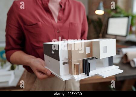Hand of young female architect or engineering designer in casualwear holding paper model of new house while standing in office Stock Photo