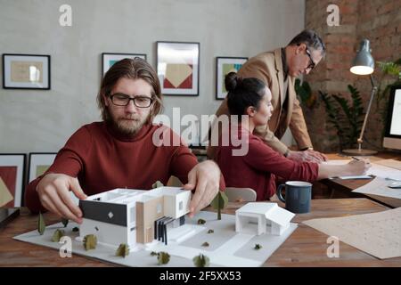 Young bearded male architect in eyeglasses and smart casualwear working over model of new house and yard against his two busy colleagues Stock Photo