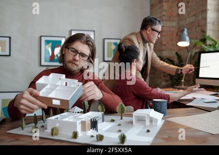 Young bearded male architect in eyeglasses holding part of new house model against his colleagues discussing sketch on computer screen Stock Photo