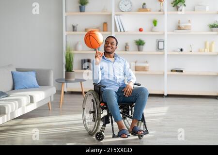 Happy disabled black guy spinning basketball on his finger, sitting in wheelchair at home Stock Photo