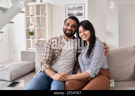 Happy indian couple hugging looking at camera sitting on couch at home. Stock Photo