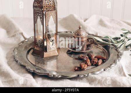 Ramadan Kareem greeting card, invitation. Bronze glowing Moroccan lantern on linen table cloth. Silver plate with dates fruit, olive branches and cup Stock Photo