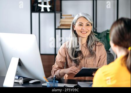 Intelligent confident influential gray-haired female asian wearing glasses, business lady, HR manager, talking to job candidate in modern office, holds and studies resume, smiling friendly Stock Photo