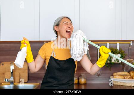 Happy funny hardworking mature gray-haired asian female cleaning worker or housewife in gloves and apron, taking a break, dancing in the kitchen with detergent and mop, singing her favorite song Stock Photo