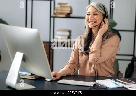 Joyful successful beautiful mature asian business woman call center manager working in office, communicating with business team via virtual conference, video call, using headset, smiling friendly Stock Photo