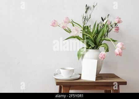 Rustic Easter still life scene. Blank greeting card mockup, cup of coffee and floral bouquet in white ceramic vase on wooden bench. Spring photo. Pink Stock Photo