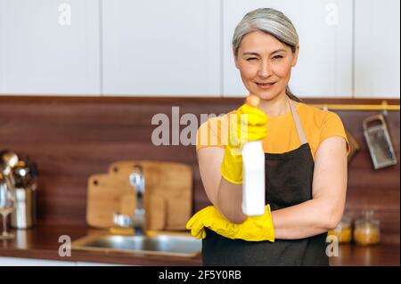 Portrait beautiful hardworking mature gray-haired Asian woman, cleaning lady or housewife in an apron and gloves, standing in the kitchen, directing detergent to camera, closing one eye, smiling Stock Photo