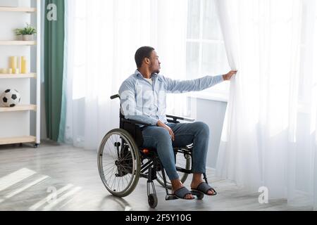 Sad disabled young man sitting on wheelchair alone at home, looking through window, full length portrait Stock Photo