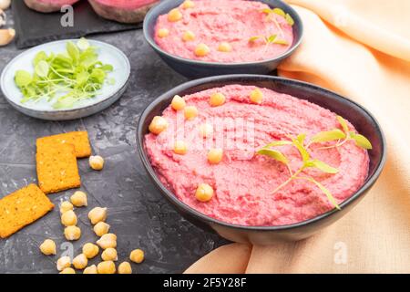 Hummus with beet and microgreen basil sprouts in ceramic bowl on a black concrete background and orange textile. Side view, close up. Stock Photo
