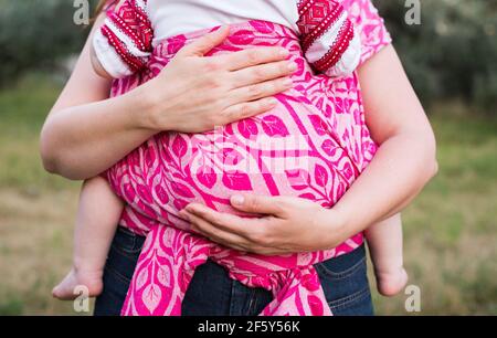 Mother hands hug baby wrapped in pink sling carrier outdoors Stock Photo