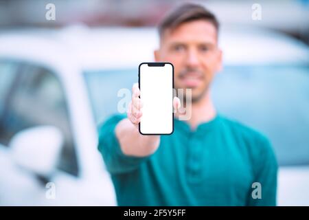 Smartphone mockup, blank screen. Young smiling man standing near white car outdoors and showing or holding his phone with blank space Stock Photo