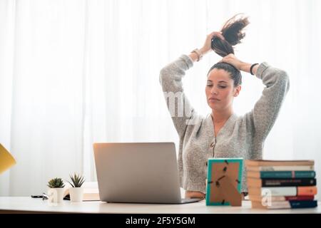 young woman working with computer at home while fixing hair Stock Photo