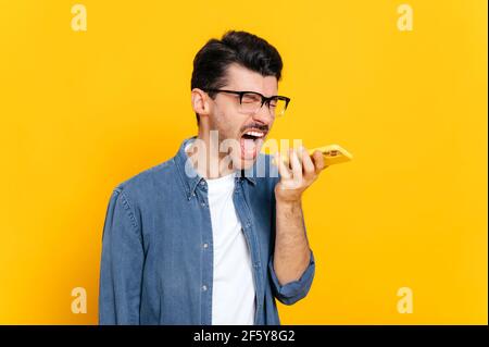Outraged angry negative caucasian stylish guy with glasses, holding smartphone in hand, shouting annoyed during phone call or recording audio message, stand against isolated orange background Stock Photo