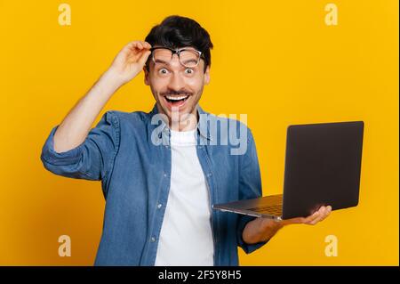 Amazed happy excited confident unshaven caucasian guy, take off glasses, holding an open laptop in his hand, looks surprised and happily at the camera while standing against isolated orange background Stock Photo