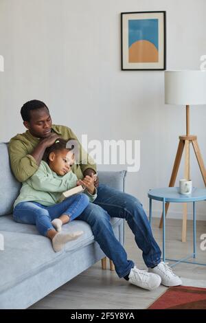 Full length portrait of loving African-American father and daughter reading while sitting on couch together in cozy home interior Stock Photo