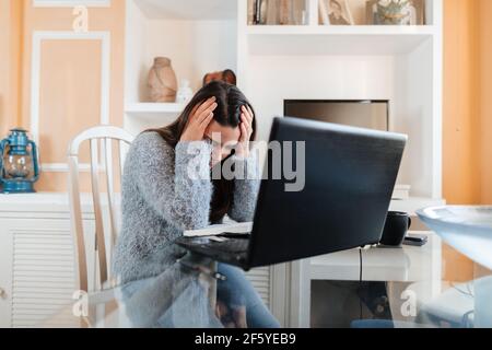 Stressed young woman wearing glasses and a sweater is sitting in the living room at home is tired of studying in online lessons Stock Photo