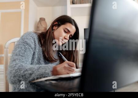 Young focused woman sitting in living room is taking notes at online class with a laptop Stock Photo