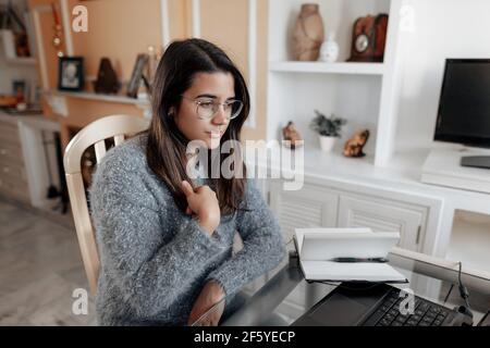 Medium shot young deaf woman wearing glasses and a sweater is using sign language to spell name while in an online video call in her living room. Stock Photo