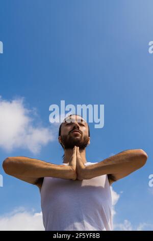 young man meditating on a rooftop. vertical photo of a man with his hair up, doing yoga on a terrace with blue sky background with clouds. Stock Photo