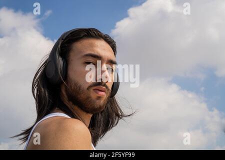 young bearded man with wireless headphones looking straight at the camera with a very serious look at the blue sky with clouds in the background. low Stock Photo