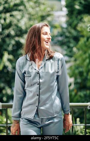 Portrait of smiling attractive young woman in silk pajamas standing on balcony of her apartment and looking away Stock Photo