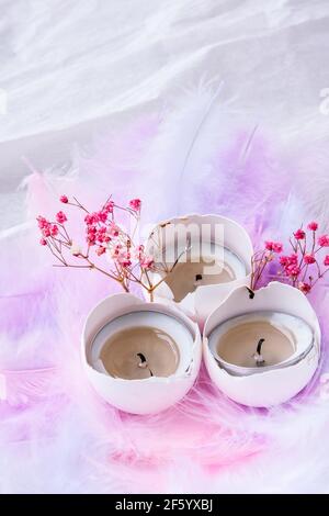 Composition of White egg shell with candle inside. Bright easter decorations with bird feathers. Pastel colors. Easter greeting card. Stock Photo