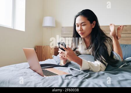 Petty young Asian woman lying on bed with opened laptop and answering text messages on smartphone Stock Photo