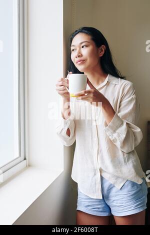 Charming pensive young Asian woman with cup of coffee looking through window Stock Photo
