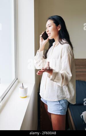 Positive young Vietnamese woman standing at window in her bedroom and talking on phone with friend Stock Photo