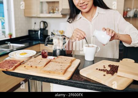 Cropped image of smiling young Asian woman pouring cream in coffee when making herself breakfast in the morning Stock Photo