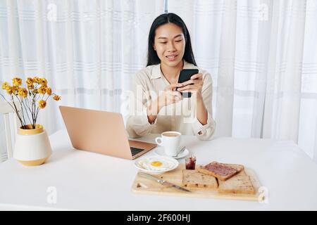 Pretty young Asian woman sitting at table, eating breakfast and checking messages on smartphone and e-mails on laptop Stock Photo