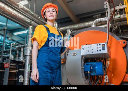 Concept of a industrial manufactory. A female smiling worker in uniform and helmet with a folder in her hand. In the background boiler room. Bottom vi Stock Photo