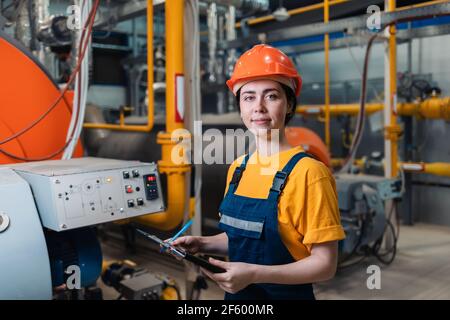 Portrait of a young smiling woman in uniform and helmet with a folder and pen in her hand. In the background, boiler equipment and pipes. Concept of i Stock Photo