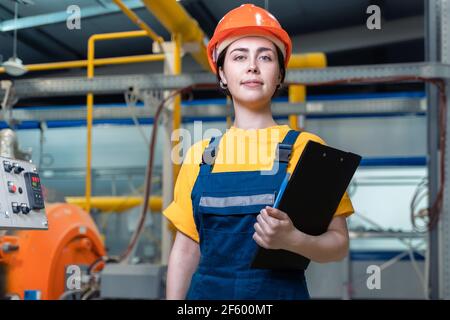 Portrait of a young smiling engineer in uniform and helmet with a folder in her hand. In the background, boiler equipment and pipes. Industrial produc Stock Photo