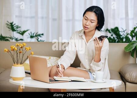 Young woman in pajamas working from home, she is listening to voice message and taking notes in planner Stock Photo