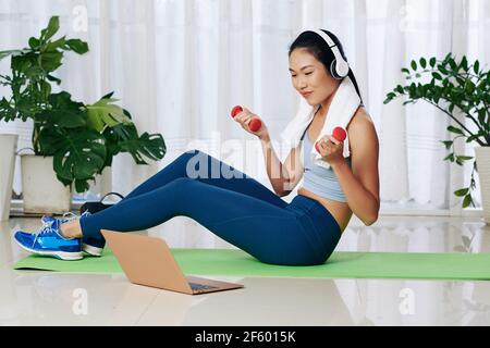 Pretty young Asian woman in headphones watching fitness video on laptop and doing exercise with dumbbells Stock Photo