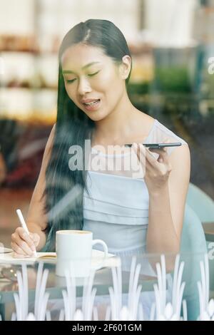 Beautiful young Asian woman working at cafe table, taking notes in planner and recording voice message for colleague Stock Photo
