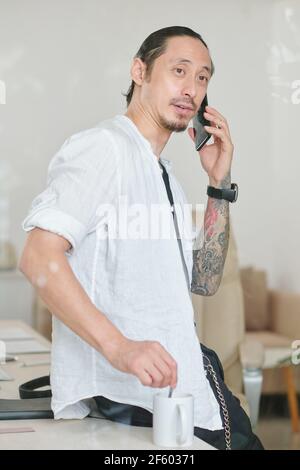Portrait of mixed-race graphic designer mixing sugar in coffee and talking on phone with client Stock Photo