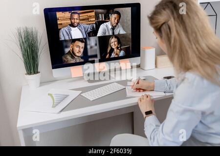 Back view of elegant female employee making notes while online conference. Successful business woman is sitting at the desk, using computer, remotely Stock Photo