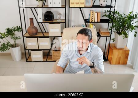 Serios frowning mixed-race entrepreneur working at desk in his home office and checking planner Stock Photo
